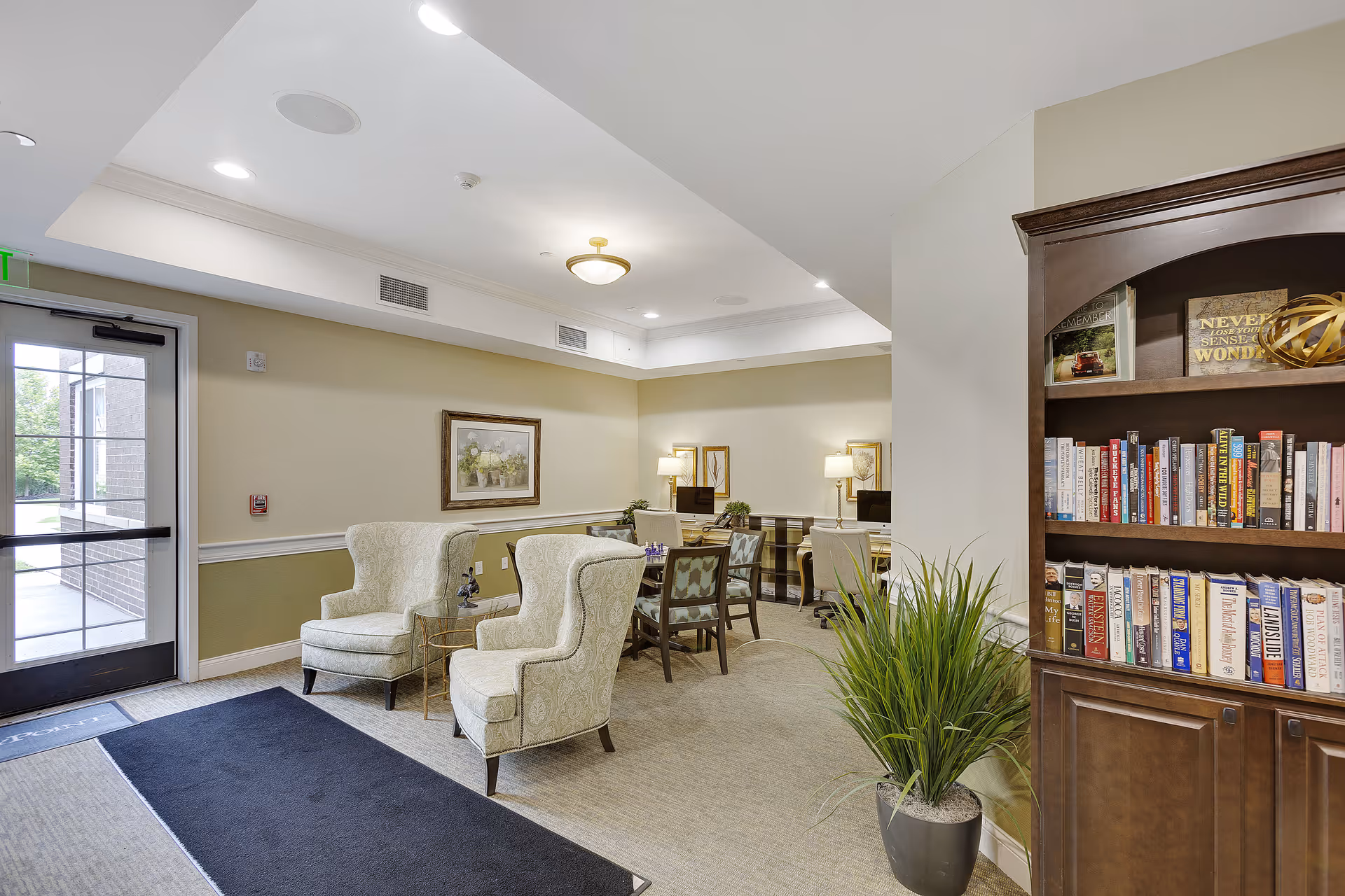A cozy interior common area with two upholstered armchairs, a small table, a wooden bookshelf filled with books, a potted plant, and a table with chairs and lamps in the background near a glass door entrance.