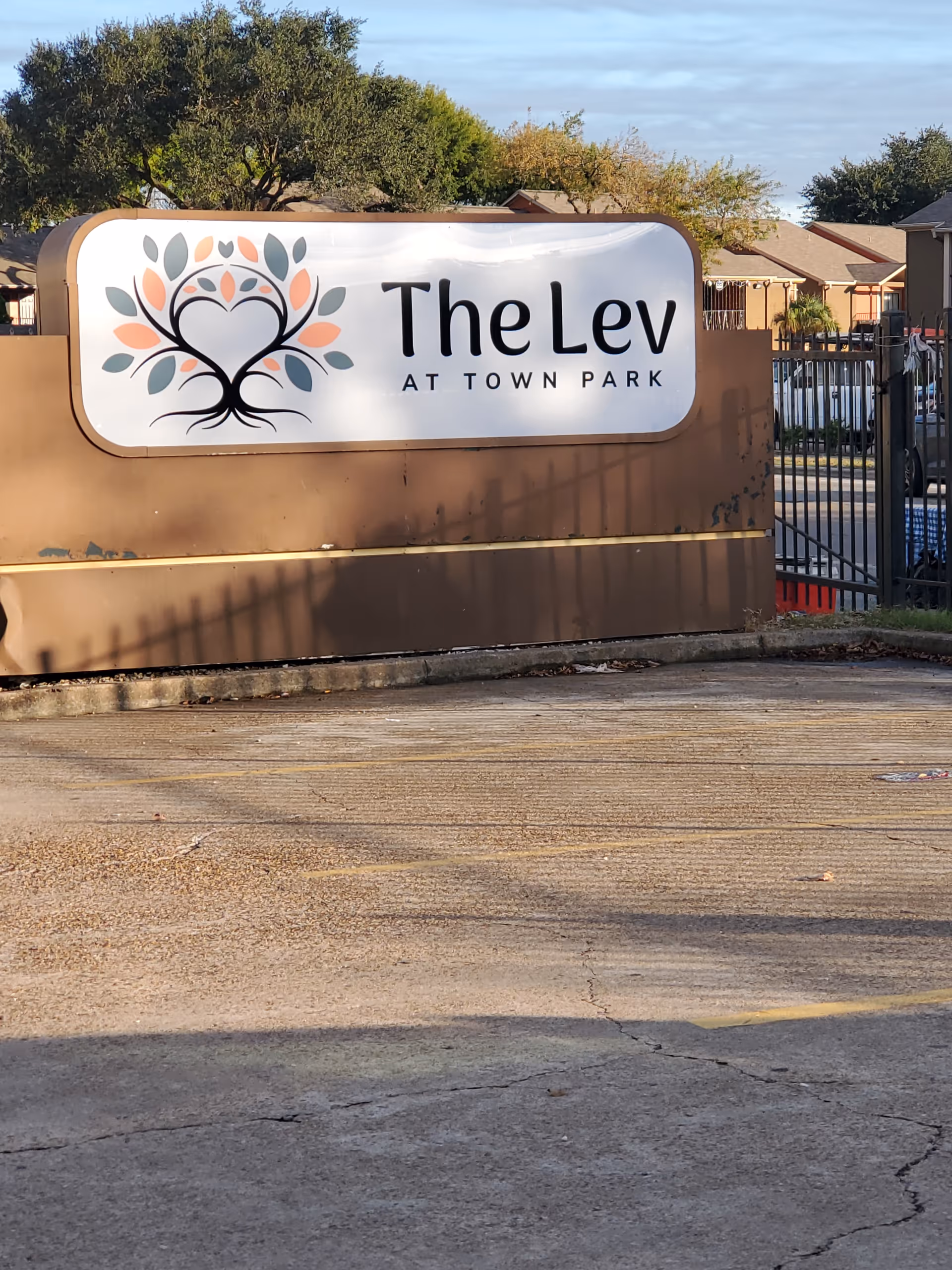 Outdoor view of a sign for The Lev at Town Park, mounted on a brown base with trees and residential buildings in the background under a partly cloudy sky.