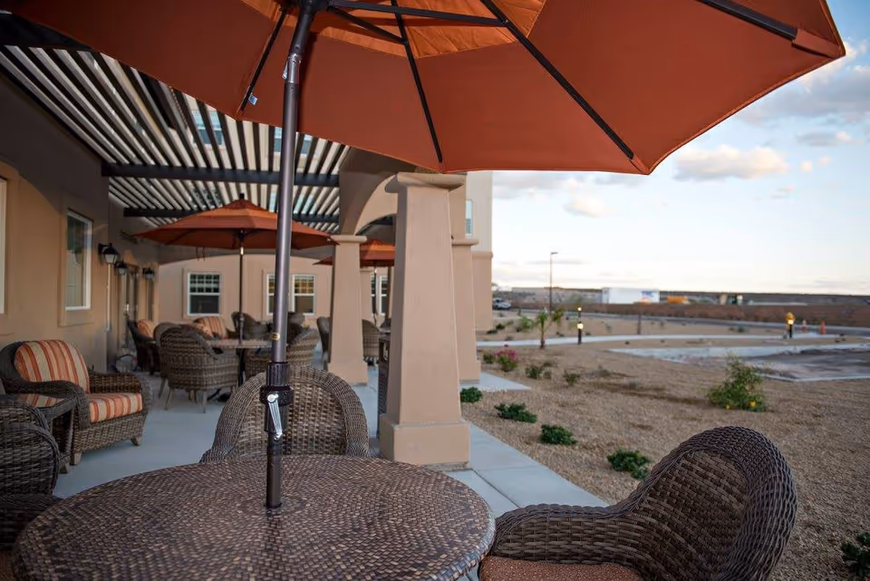 Outdoor patio area at Joshua Springs Senior Living with round wicker tables and chairs, each table shaded by large orange umbrellas. The patio is covered partially by a pergola attached to the building, with beige walls and windows. The surrounding area has sparse landscaping with small bushes and a clear sky in the background.