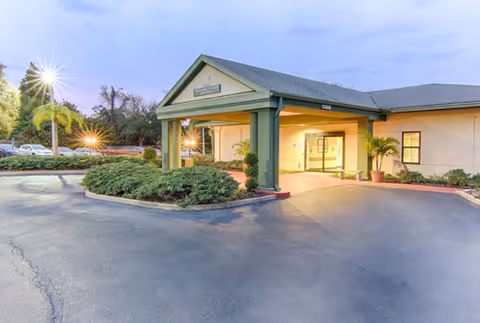 Exterior view of Solaris HealthCare Bayonet Point building entrance with a covered driveway, green columns, and surrounding landscaping under a twilight sky.