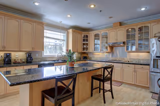 Bright and spacious kitchen with light wood cabinets, granite countertops, a large island with two chairs, a window above the sink, and various kitchen appliances including a coffee maker and refrigerator.