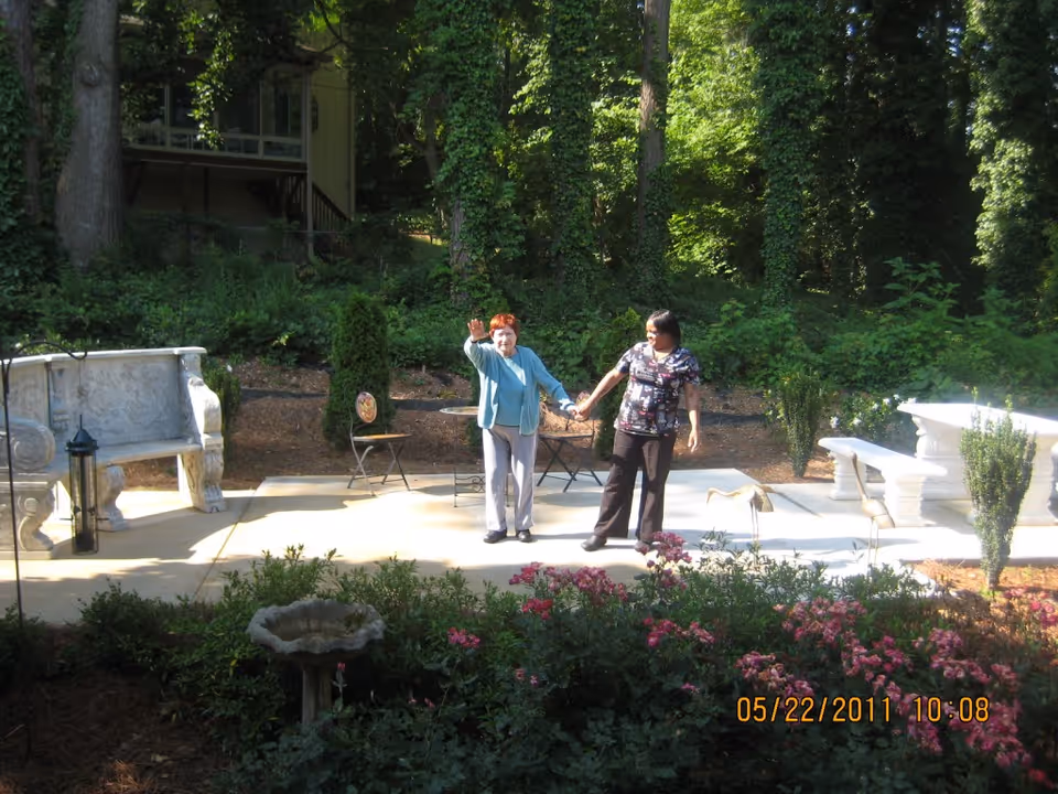 An elderly woman and a caregiver holding hands and walking together on a paved patio surrounded by greenery and flowers. The patio has stone benches and small tables, with a wooded area in the background.