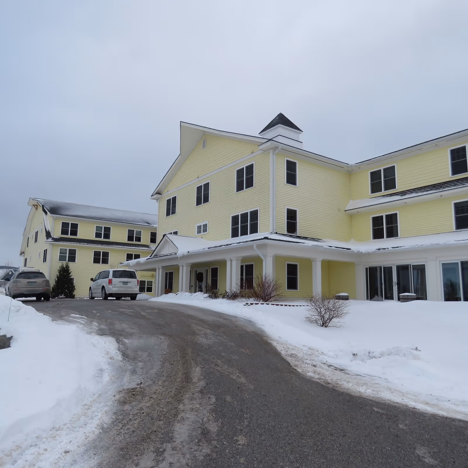 Exterior view of a large yellow senior living facility building with multiple windows and a covered entrance, surrounded by snow and a driveway with parked vehicles.