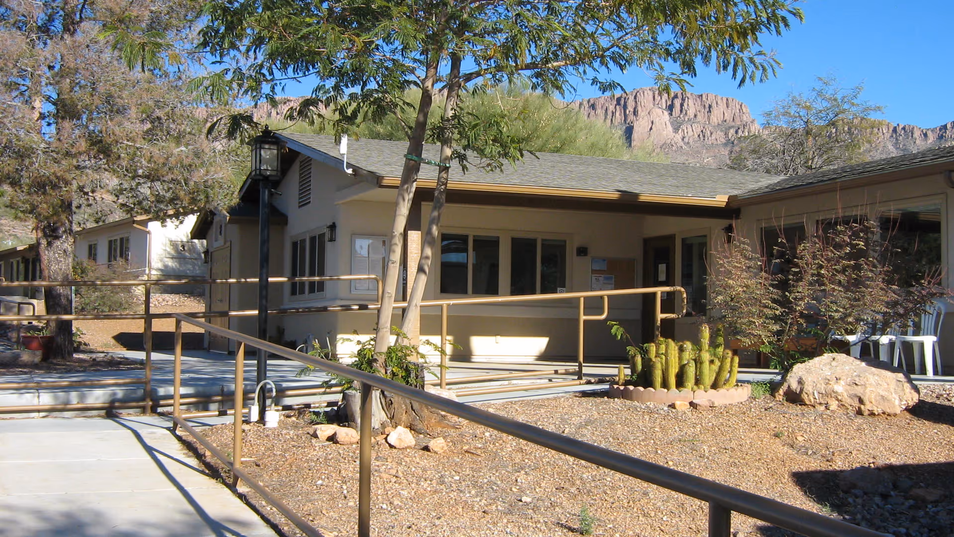 Exterior view of a single-story building with beige walls and a gray roof, surrounded by desert landscaping including cacti and rocks. There is a metal handrail along a concrete pathway leading to the entrance. Trees and a mountain are visible in the background under a clear blue sky.