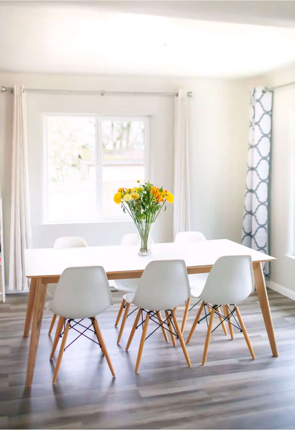 A bright dining area with a table and white chairs, featuring a vase of yellow flowers.