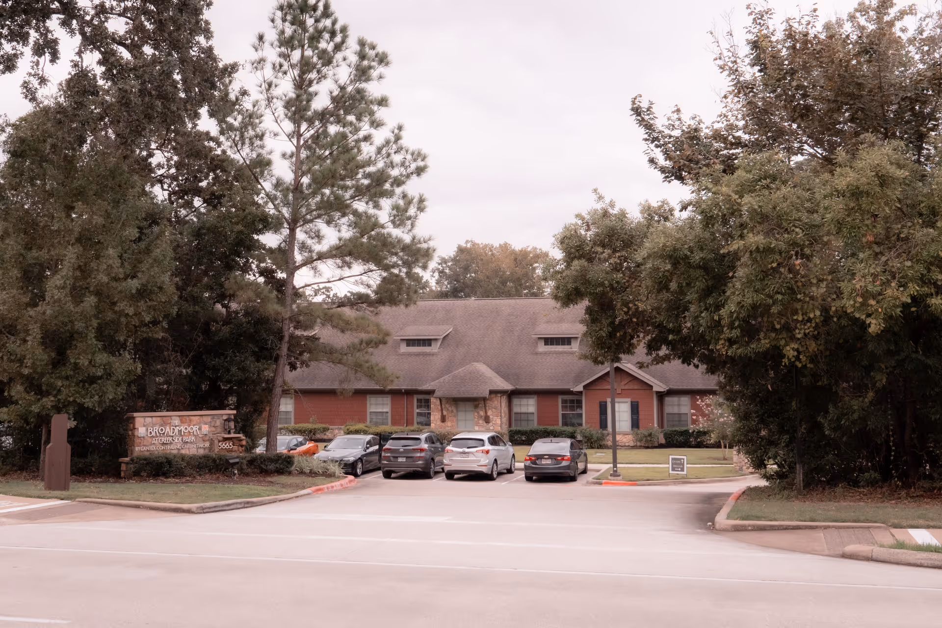 Exterior view of The Broadmoor At Creekside Park building with a parking lot in front containing several cars. The building is surrounded by trees and greenery, and a stone sign with the facility name is visible on the left side near the entrance.