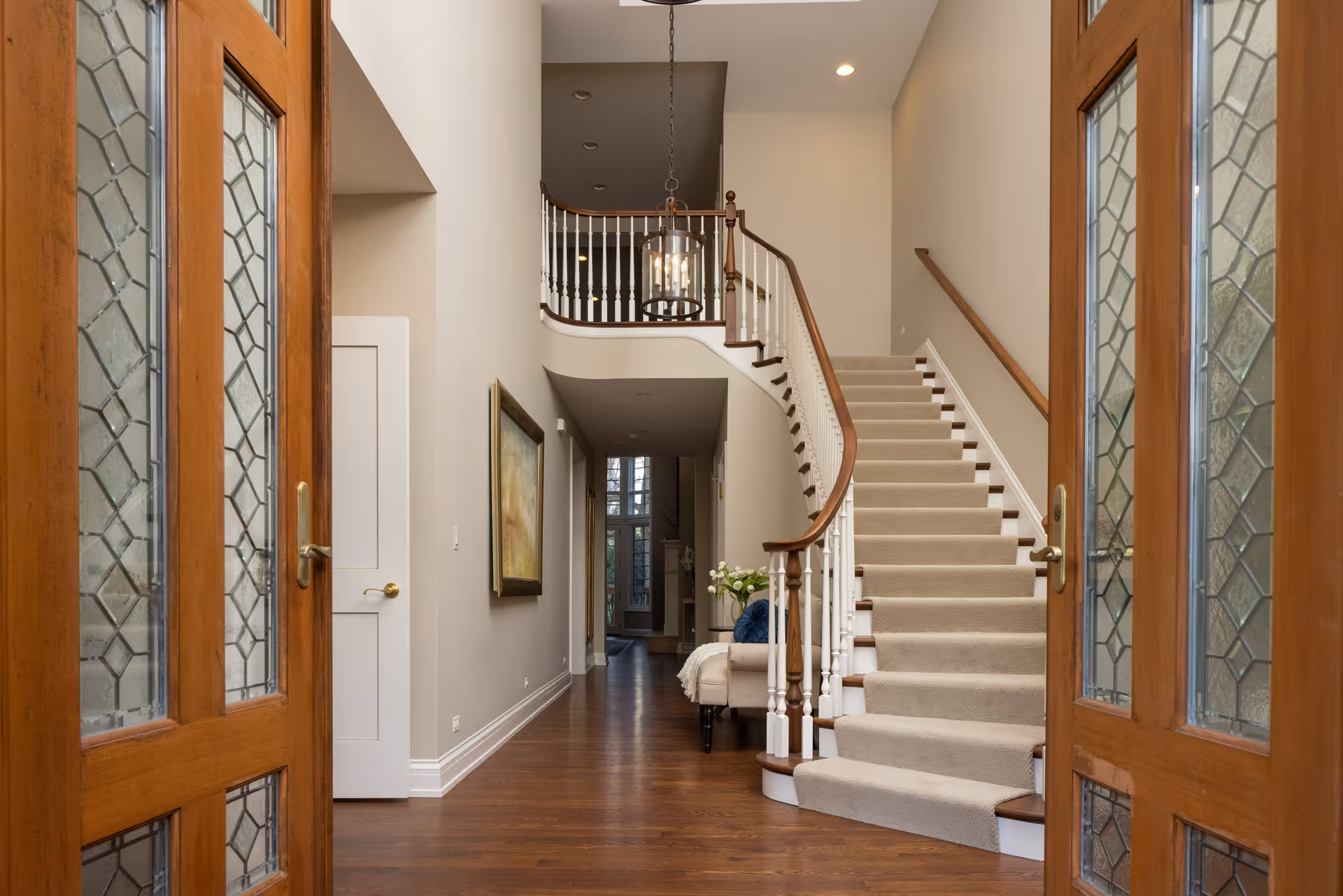 View through wooden double doors with decorative glass panels into a hallway featuring a carpeted staircase with wooden handrails and white balusters. The hallway has hardwood floors, a painting on the left wall, a small bench with cushions and flowers near the staircase, and a hanging light fixture above. The space is well-lit with recessed ceiling lights.