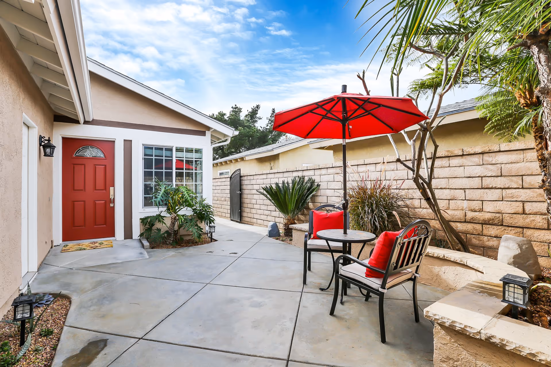 Outdoor patio area with a red door on the left, a window, and a small garden with plants. There is a round table with two chairs featuring red cushions and a large red umbrella providing shade. The patio is surrounded by a beige brick wall and some greenery including palm trees and shrubs.