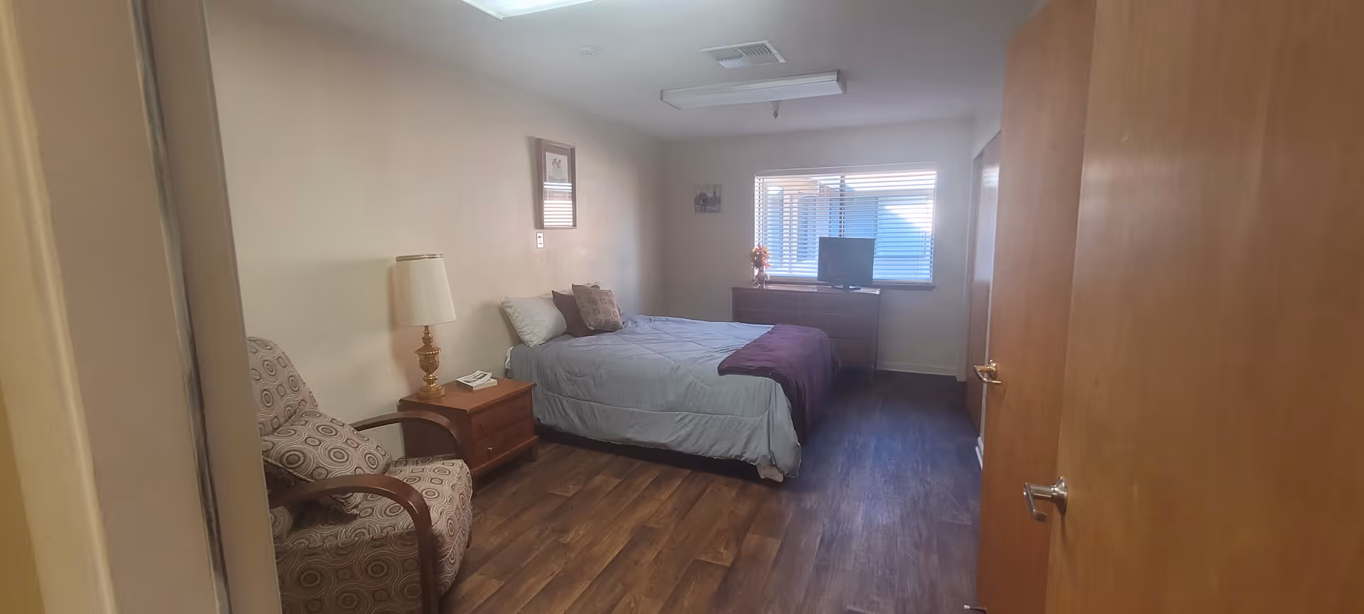 Furnished bedroom with a bed, nightstand and lamp, upholstered chair, dresser with TV, and a window.