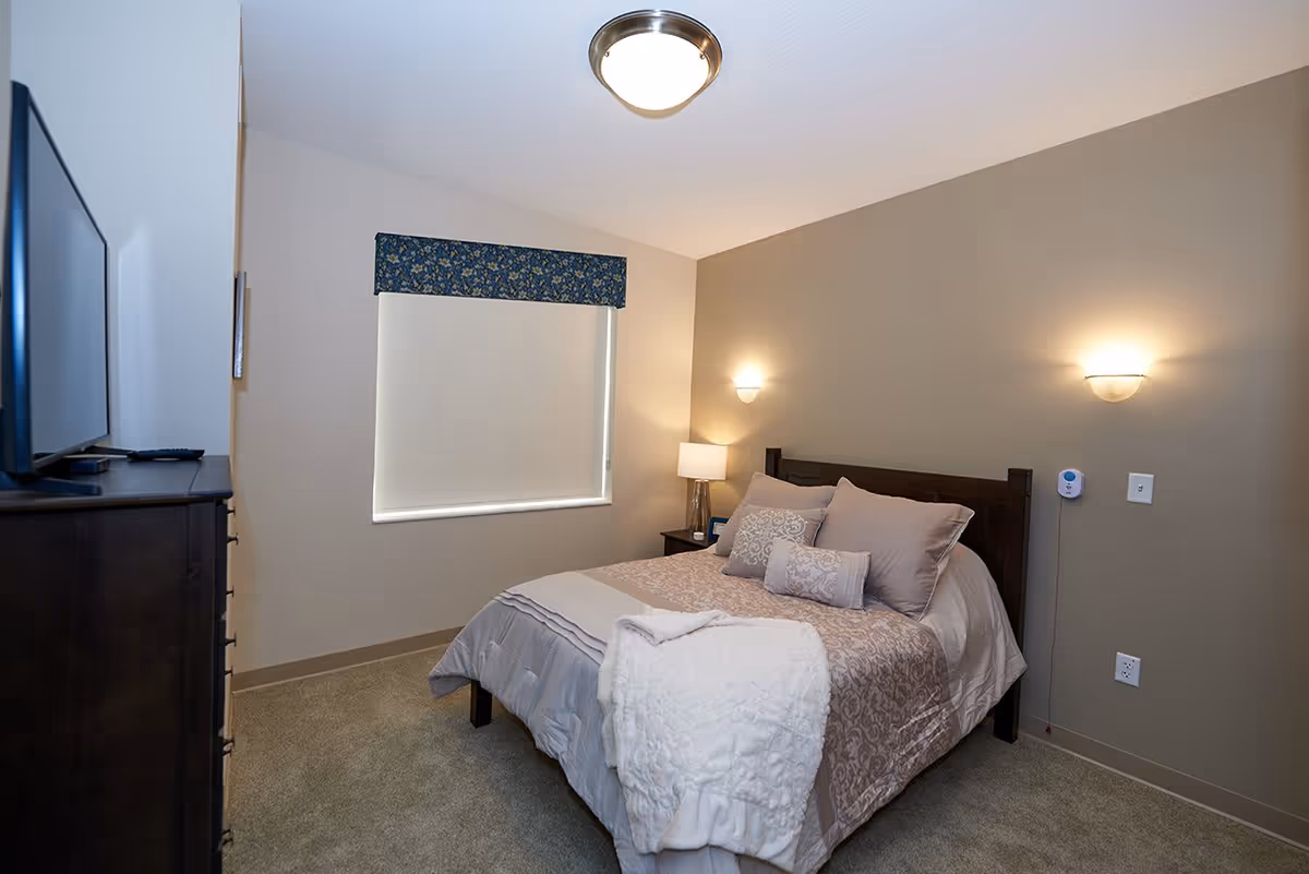 A cozy bedroom with a neatly made bed featuring multiple pillows and a folded white blanket at the foot. The room has beige walls, a window with a floral valance and a closed blind, a dark wooden dresser with a TV on top, and soft lighting from wall sconces and a bedside lamp.