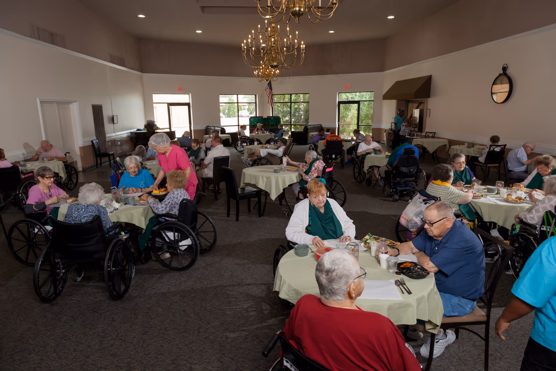 A dining room in Autumn Woods Health Care Facility with multiple elderly residents seated at round tables covered with light green tablecloths. Many residents are in wheelchairs, eating or being assisted by staff members. The room has large windows letting in natural light, a chandelier hanging from the ceiling, and a warm, communal atmosphere.