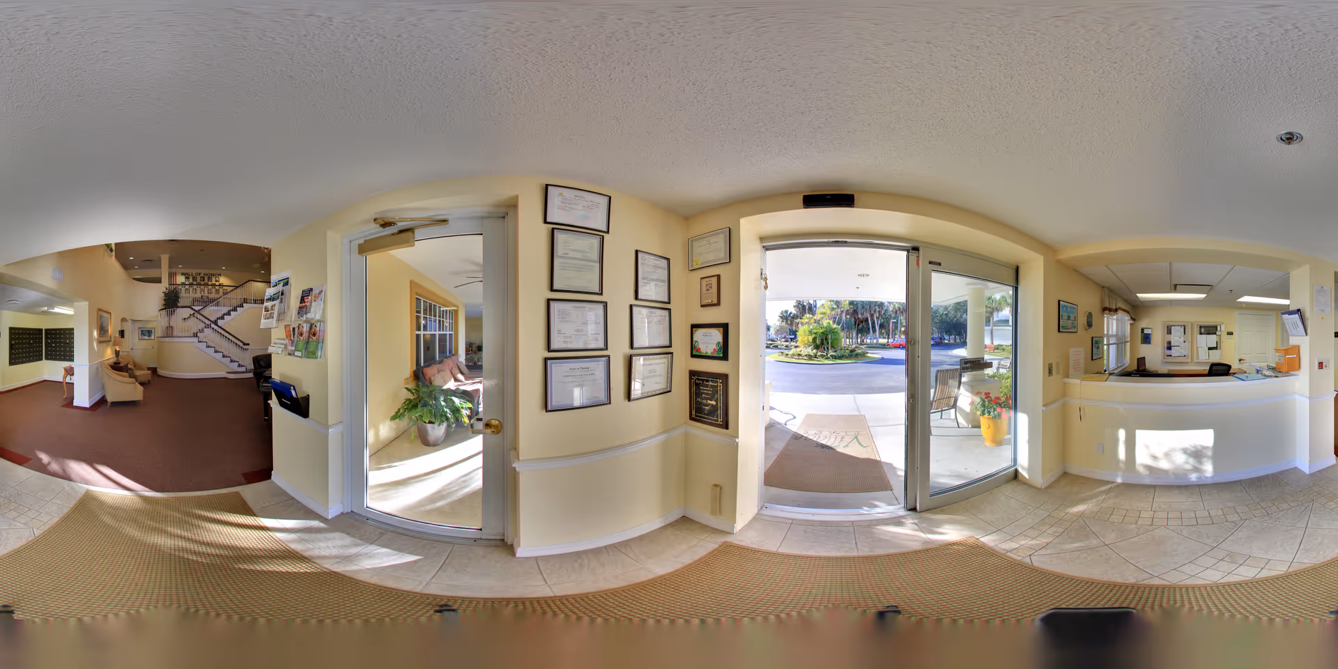 Wide panoramic view of the entrance lobby of Truewood by Merrill, Charlotte Center, showing a reception desk on the right, glass doors leading outside in the center, and a hallway with seating and stairs on the left. The walls are decorated with framed certificates and artwork, and the floor is tiled near the entrance with carpet further inside.