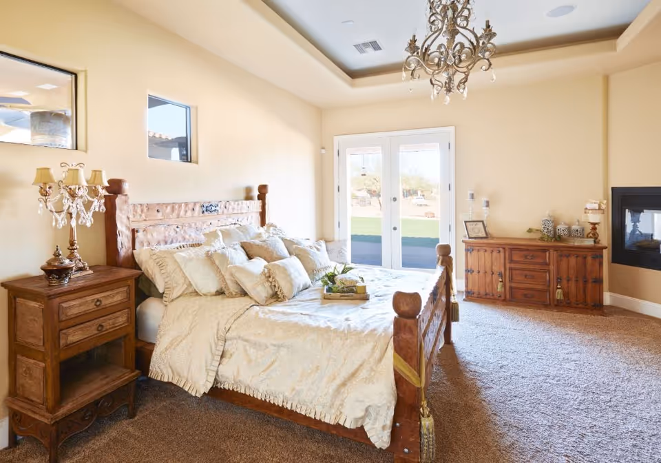 A bright and spacious bedroom with a large wooden bed adorned with multiple cream-colored pillows and a matching bedspread. There is a wooden nightstand with an ornate lamp on the left side of the bed and a wooden dresser with decorative items on the right side of the room. French doors open to an outdoor area, and a chandelier hangs from the ceiling. The room has beige walls and carpeted flooring.