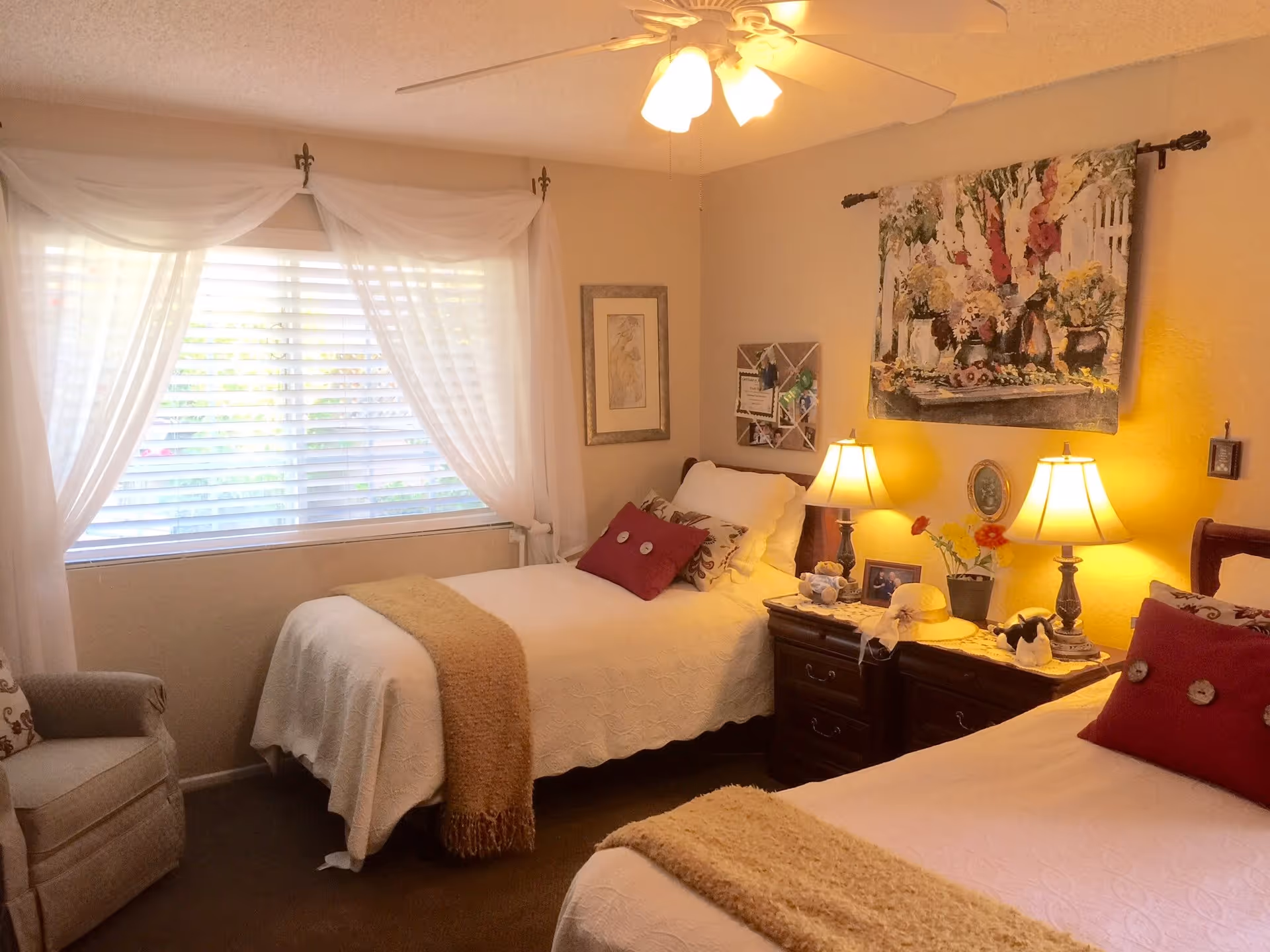 Warmly lit bedroom with two twin beds, a shared nightstand with lamps, an armchair, window with sheer curtains, and wall art.