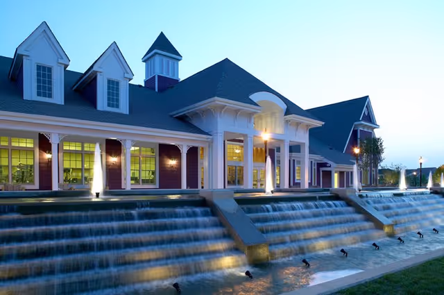 Exterior view of a large building with a steep roof and multiple dormer windows, illuminated at dusk. In front of the building is a tiered water feature with cascading waterfalls and lights, surrounded by a well-maintained lawn and trees.