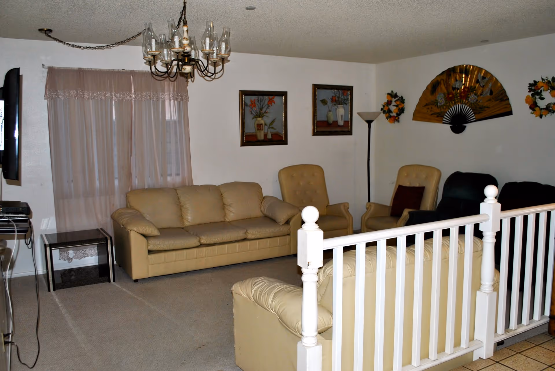 A living room with beige leather sofas and armchairs arranged around a white railing. The room has a chandelier, two framed floral paintings on the wall, a large decorative fan, and two floral wreaths. There is a window with sheer curtains and a floor lamp in the corner.