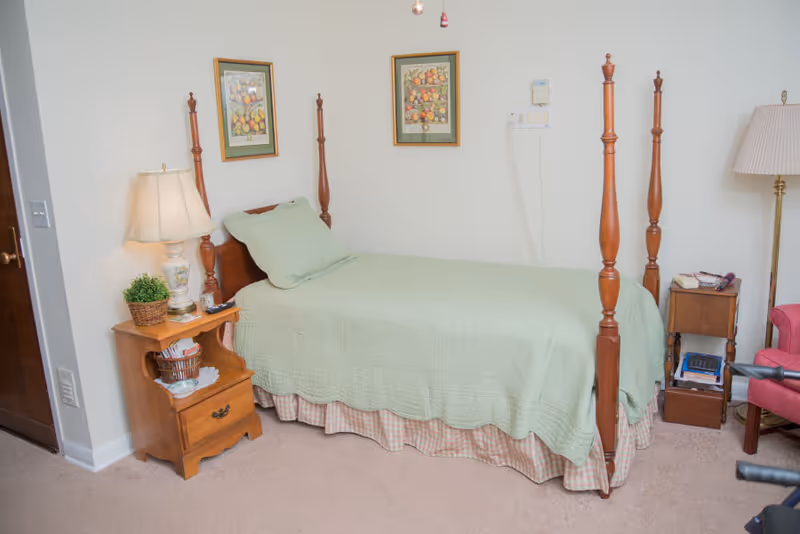 A bedroom with a wooden four-poster bed covered with a light green quilt and matching pillow. There is a wooden nightstand with a lamp, a small plant, and some items on it next to the bed. On the wall above the bed are two framed pictures of fruit. Another small wooden table with a lamp and some books is next to a pink armchair on the right side of the room. The carpet is beige and the walls are white.