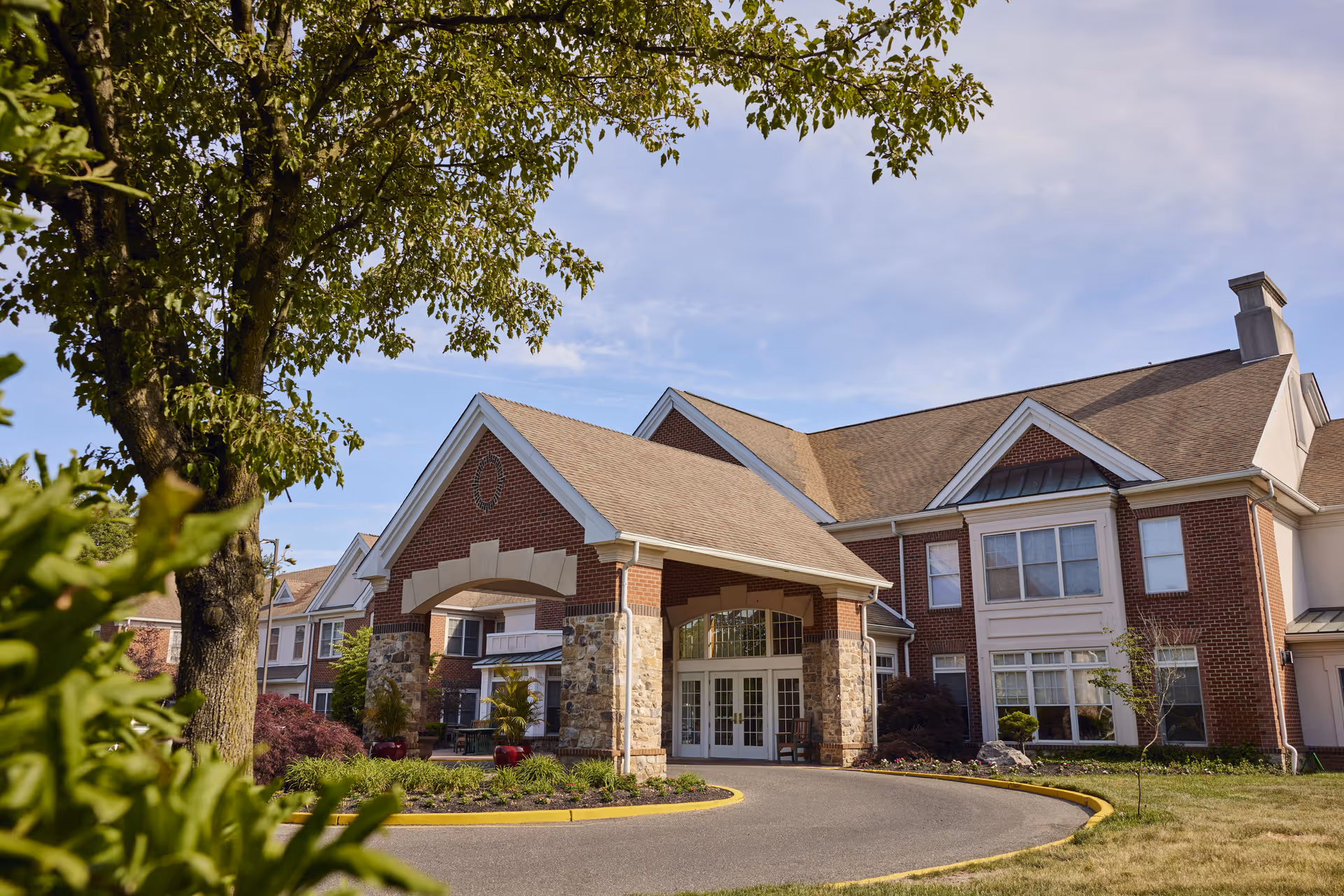 Exterior view of a senior living facility building with a covered entrance supported by stone pillars, surrounded by landscaped greenery and trees under a clear blue sky.