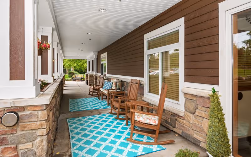 Covered outdoor porch area with wooden rocking chairs placed on blue patterned rugs. The porch has brown siding with stone accents and white trim. There are potted plants and flowers along the left side, and a bench is visible at the far end near green trees and grass.
