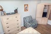 A corner of a bedroom in a retirement community featuring a light wood dresser with four drawers, a patterned armchair with blue and gray tones, a wooden entertainment cabinet with a TV inside, and a framed picture of a bird on the wall. The floor is wood, and part of a bed with a light-colored bedspread is visible in the foreground.