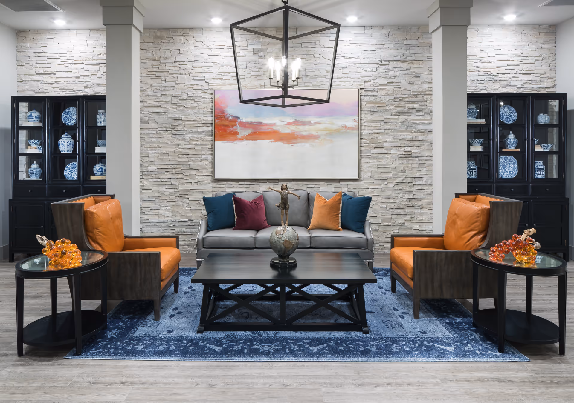 Comfortable lobby-style lounge with a gray sofa flanked by orange leather chairs, a central coffee table, decorative cabinets, and abstract wall art on a textured stone wall.
