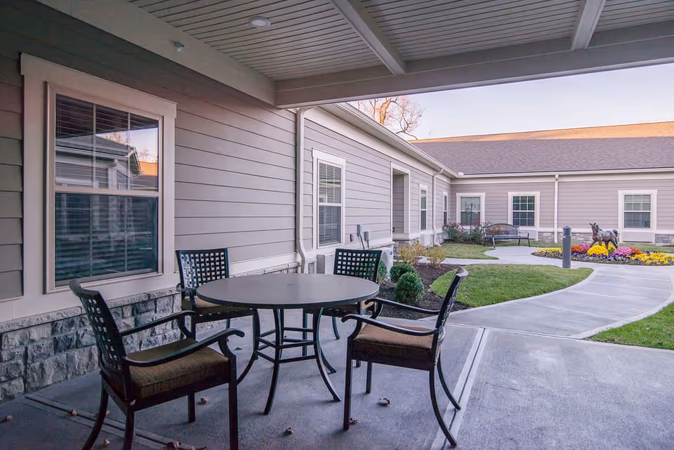Outdoor patio area at a senior living facility with a round table and four chairs under a covered porch. The patio overlooks a landscaped garden with a curved concrete walkway, green grass, colorful flowers, a bench, and a small horse statue. The building exterior is light gray with white trim and multiple windows.