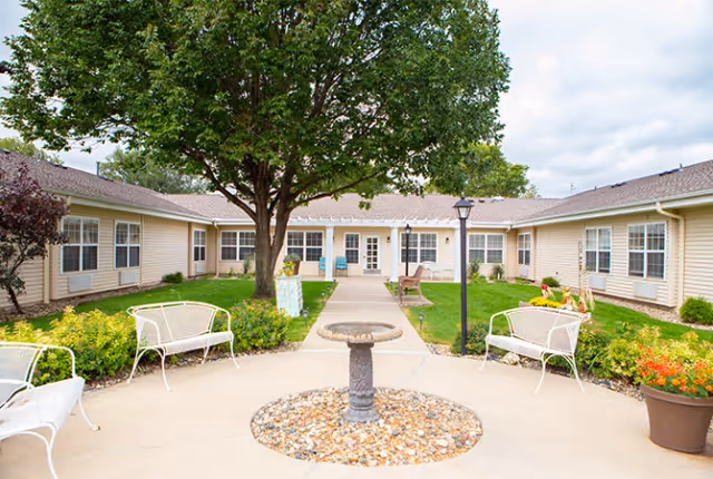 Outdoor courtyard area of a senior living facility with a central stone birdbath surrounded by a circular bed of small rocks, white metal benches, green grass, shrubs, and a large tree. The courtyard is enclosed by a single-story beige building with multiple windows and a central entrance door. A lamppost is also visible near the birdbath.