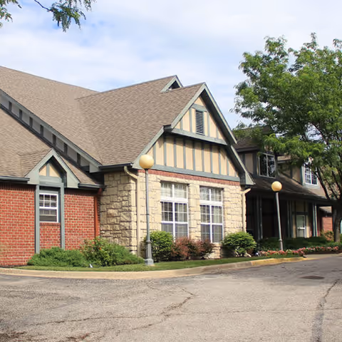 Exterior view of a senior living facility building with a combination of brick and stone walls, large windows, and a sloped roof. There are two lamp posts with round lights along the driveway, and some greenery including bushes and trees around the building.