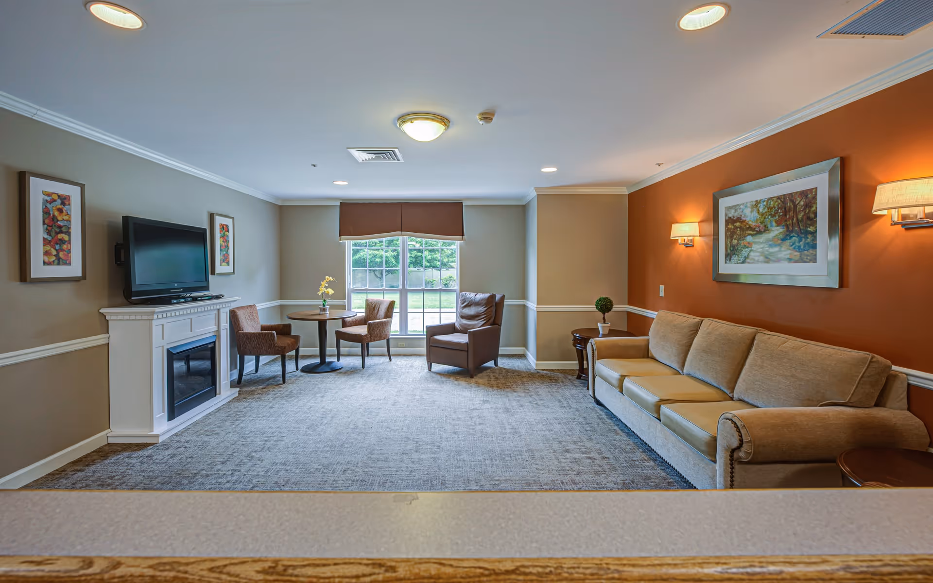 A cozy living room with a beige sofa against an orange accent wall, two wall-mounted lamps, a framed landscape painting, a small round table with two chairs near a large window, a leather armchair, a white fireplace with a flat-screen TV mounted above it, and two framed floral artworks on the adjacent wall.