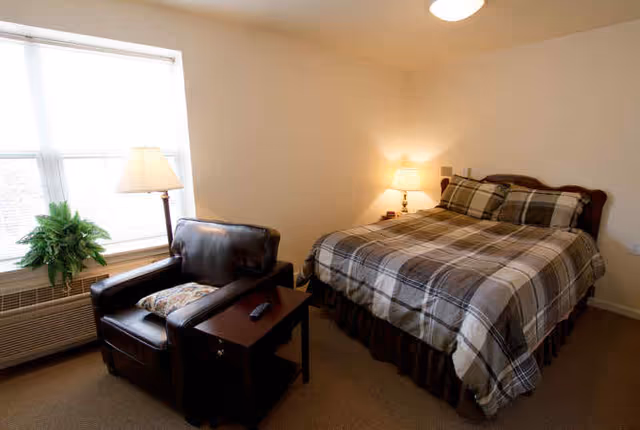 A neatly furnished bedroom with a bed, armchair, side table, and window.