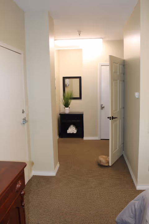 Interior hallway of a senior living facility with beige walls and carpeted floor. There is a small black cabinet with rolled white towels and a potted plant underneath a wall mirror. Two doors are visible, one partially open with a doorstop, and a piece of wooden furniture is partially visible on the left side.