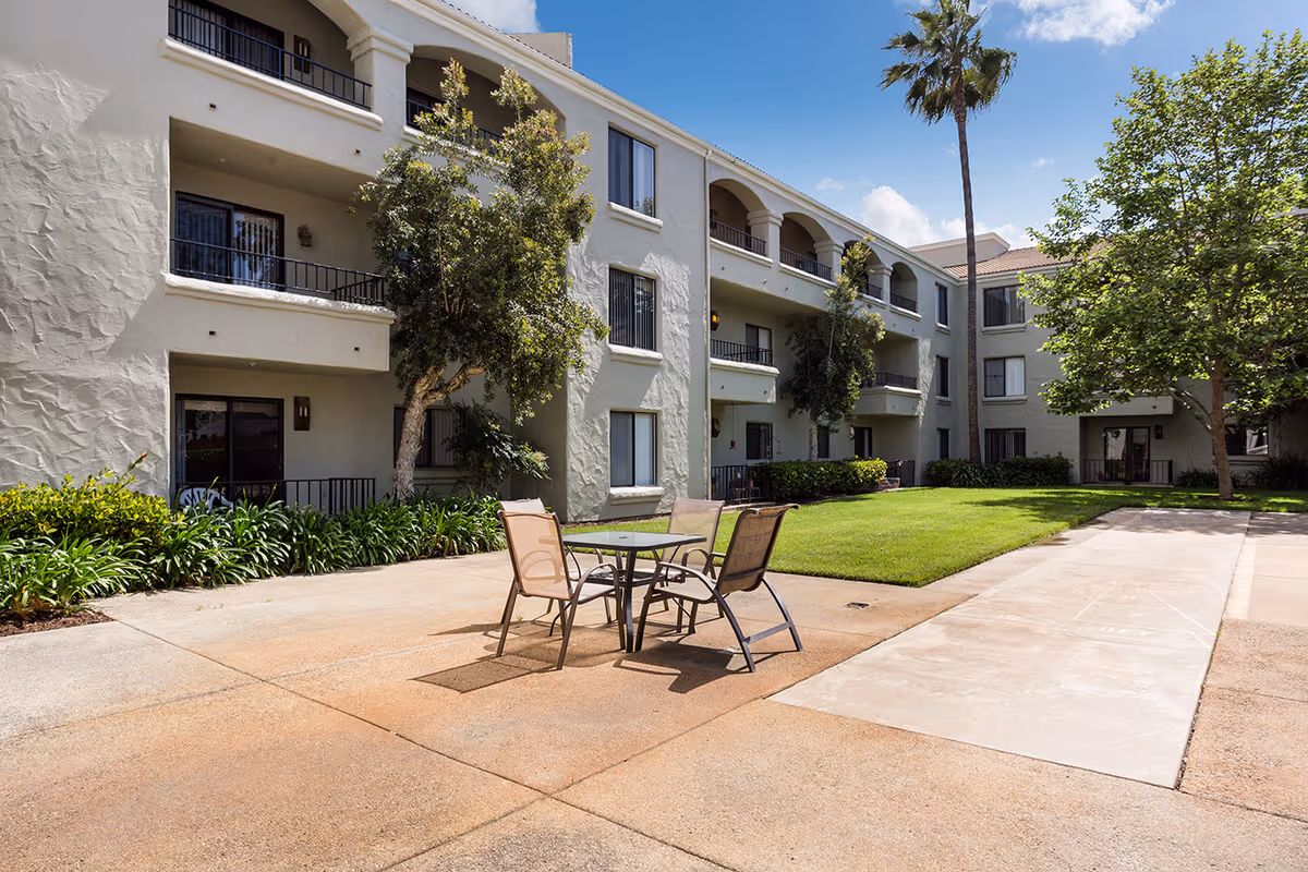 Courtyard of a three-story senior living building with a patio table and chairs, balconies, lawn, and palm trees.