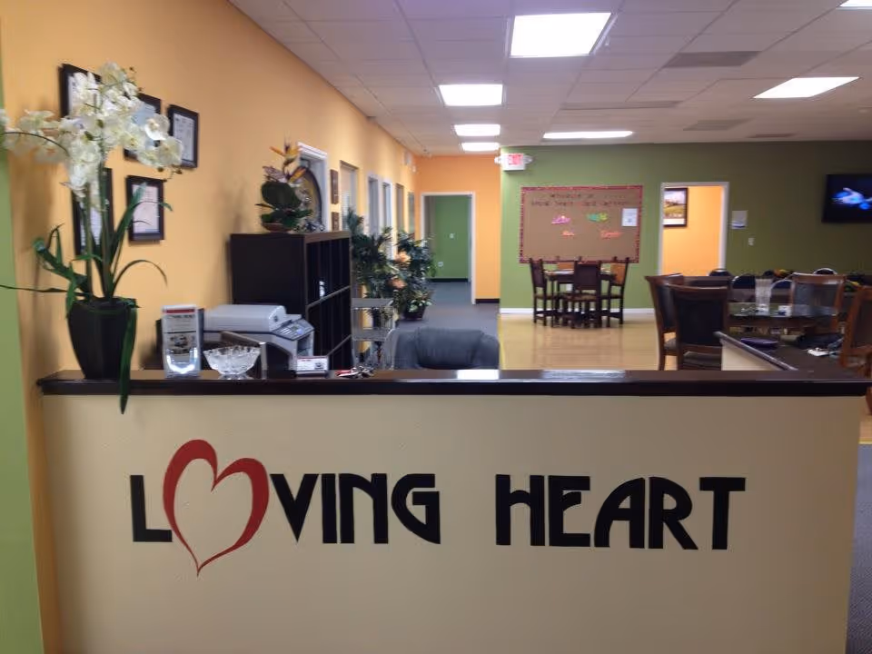 Reception desk with a 'Loving Heart' logo overlooking a brightly colored common area with tables, plants, and seating.