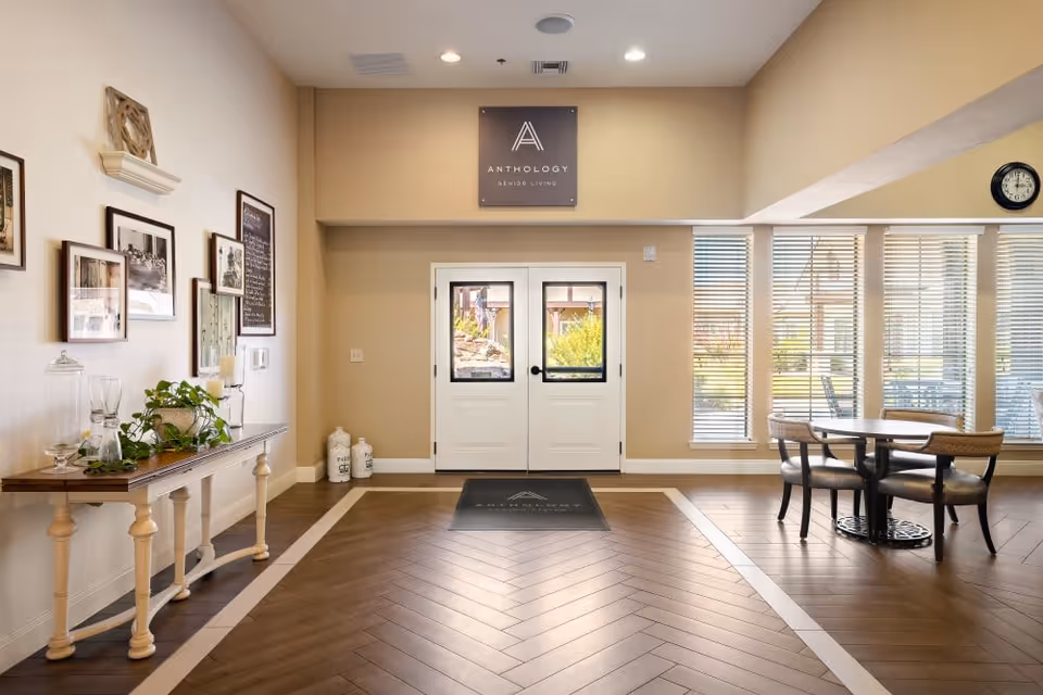 Bright senior living lobby with double entry doors centered, a decorative console table on the left, and a seating area by large windows on the right.