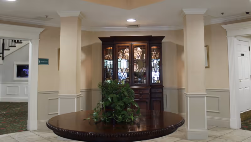 Interior view of a senior living facility lobby area with a round wooden table topped with a green leafy plant in the center. Behind the table is a wooden cabinet with glass doors displaying decorative items. The walls are beige with white trim, and there are two doorways visible, one leading to a carpeted area with stairs and the other to a hallway with a white door.