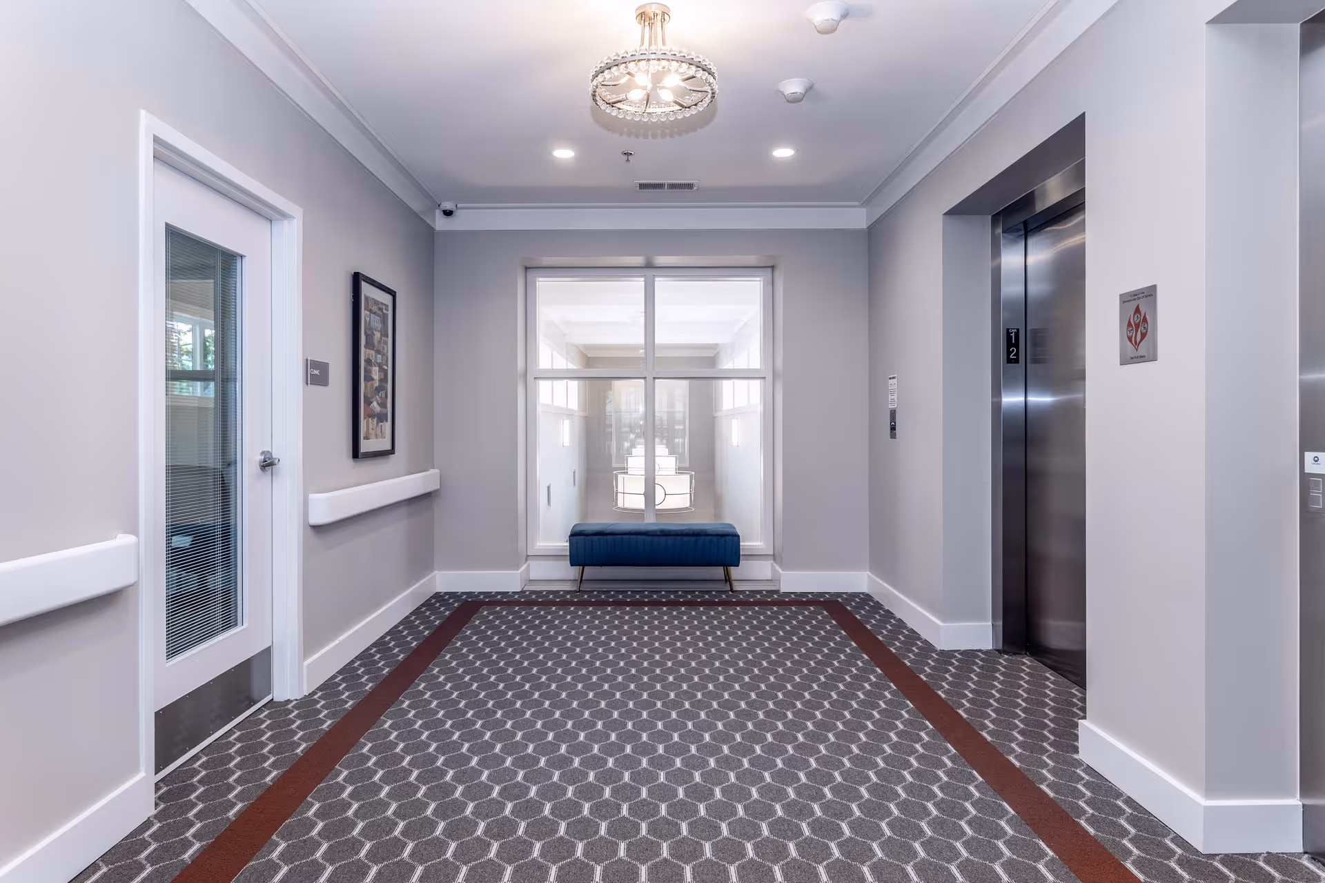 A clean and modern hallway area in a senior living facility with a patterned carpet, a blue bench in front of a large window, a chandelier light fixture on the ceiling, an elevator with closed doors on the right, and a door with a glass panel on the left.