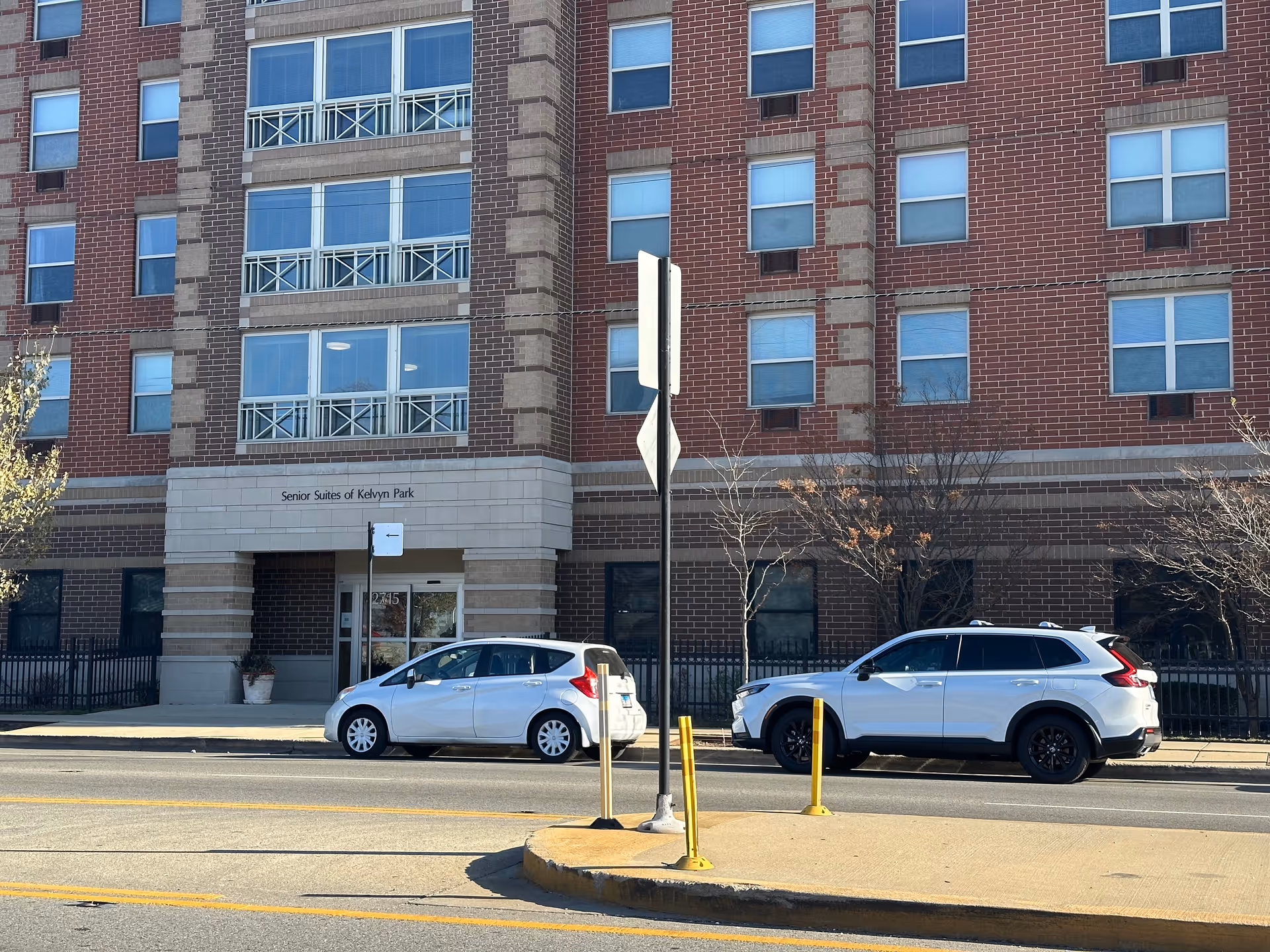 Exterior view of a multi-story brick building with large windows and a sign above the entrance that reads 'Senior Suites of Kelvyn Park'. Two white cars are parked on the street in front of the building, and there are some leafless trees along the sidewalk.