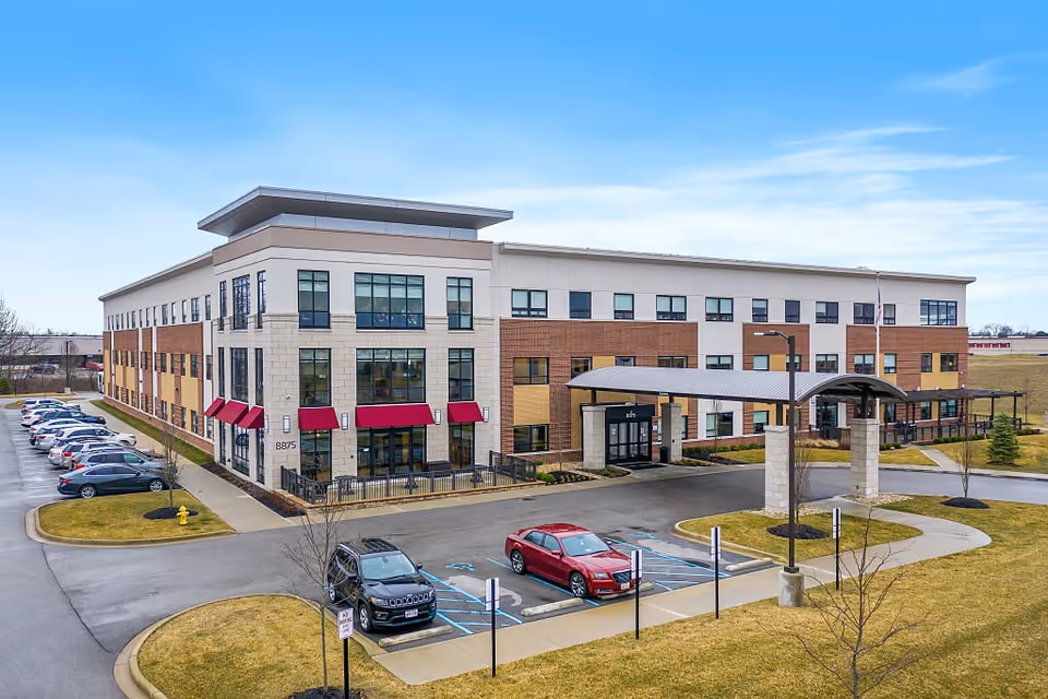 Exterior view of The Avalon of Lewis Center, a modern three-story senior living facility with large windows, red awnings, and a covered entrance. There are parked cars in the parking lot and a well-maintained lawn surrounding the building under a clear blue sky.