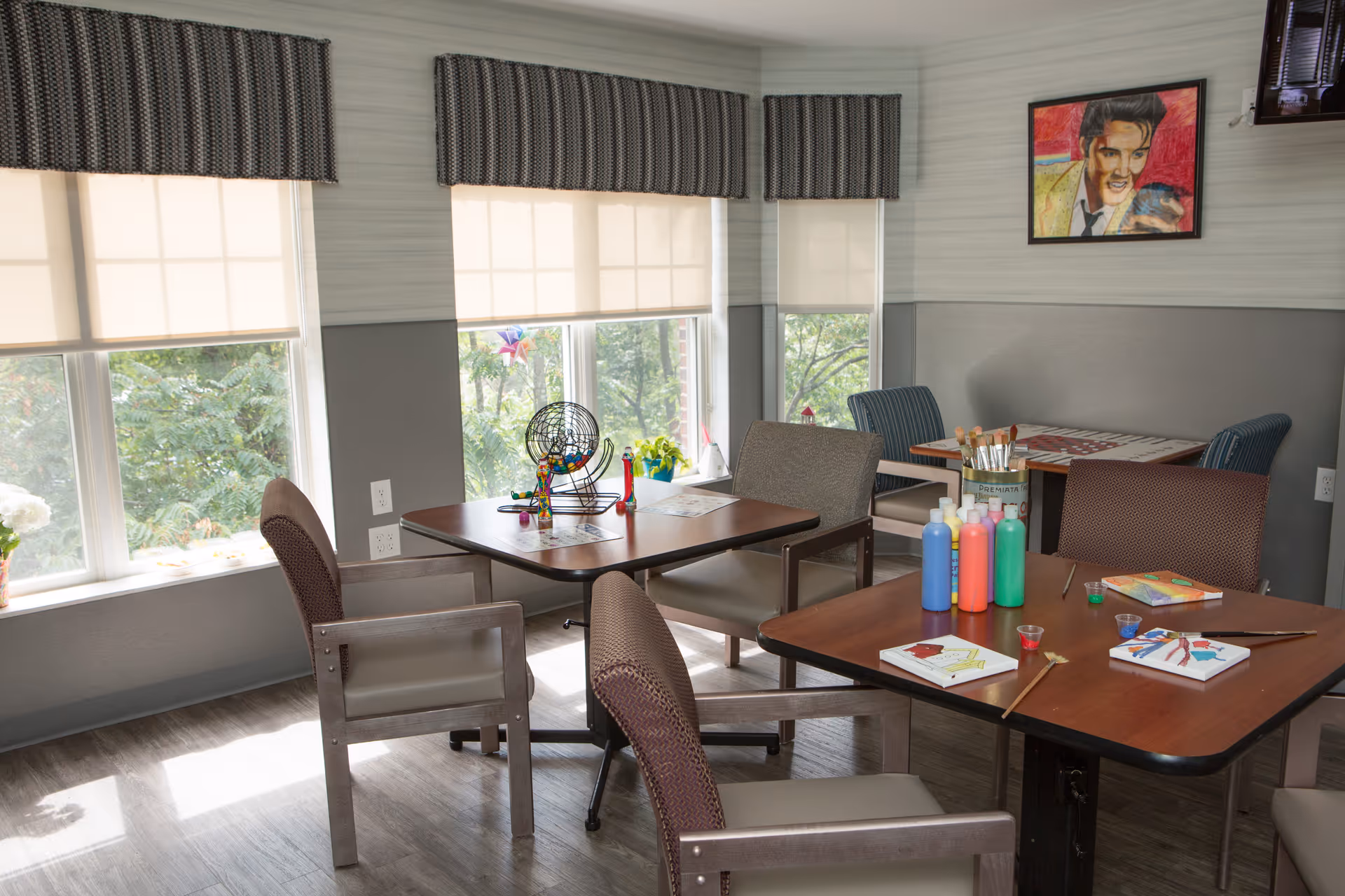 A bright activity room with large windows covered by white roller shades and patterned valances. The room contains several tables and chairs arranged for group activities. On one table, there are colorful bottles of paint, paintbrushes, and small canvases with paintings in progress. Another table has a bingo cage and cards. A framed colorful portrait hangs on the wall. The floor is wood-patterned, and the walls are painted in two tones of gray.