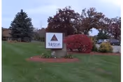 Outdoor view of a grassy area with a sign that reads 'Metron' mounted on a stone base, surrounded by landscaped bushes and trees with autumn foliage.