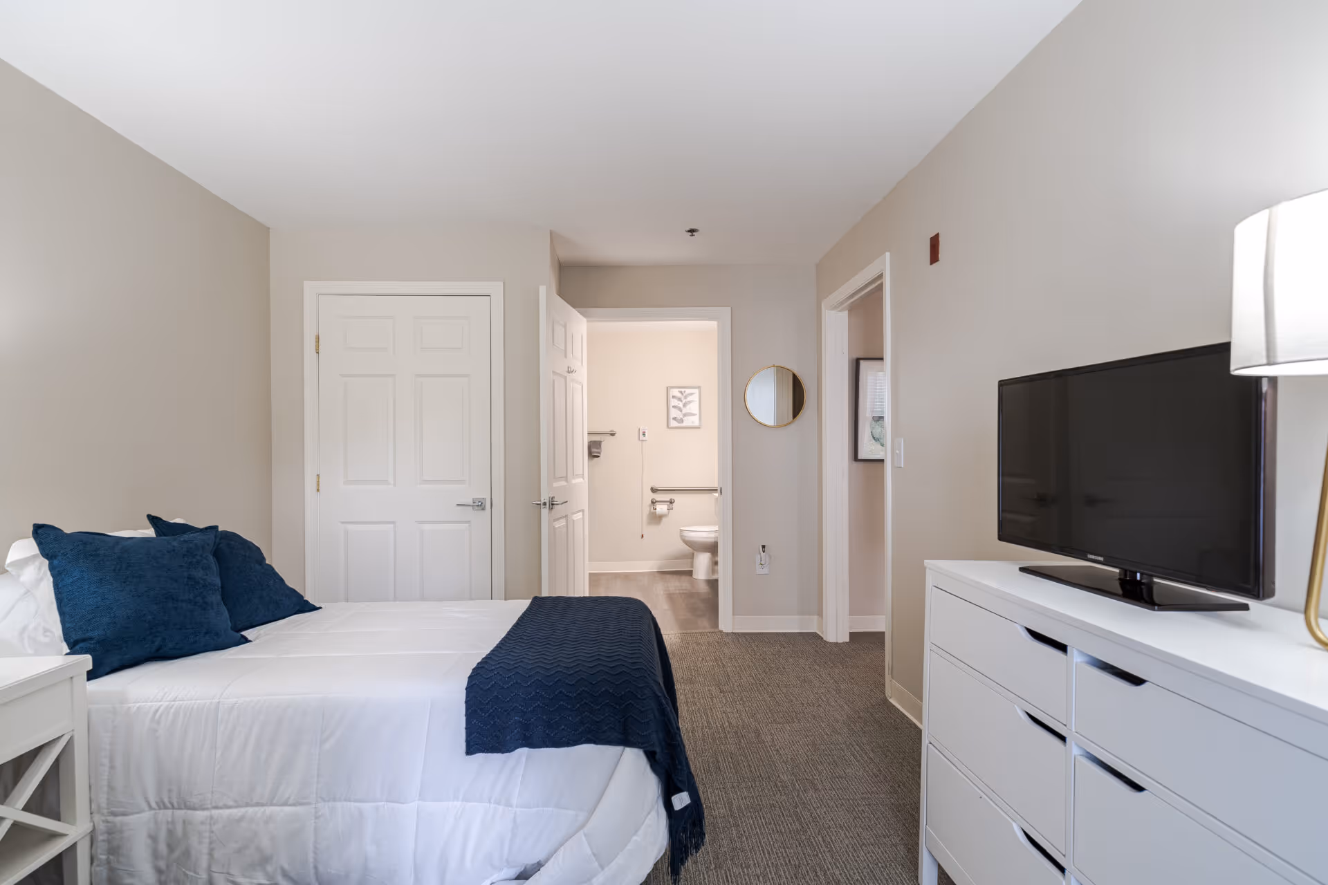 A neatly arranged bedroom with a white bedspread and dark blue pillows and throw blanket. There is a white nightstand beside the bed and a white dresser with a flat-screen TV on top. The room has beige walls and carpeted flooring. An open door reveals a bathroom with a toilet and grab bars, and another door leads to an adjacent room.