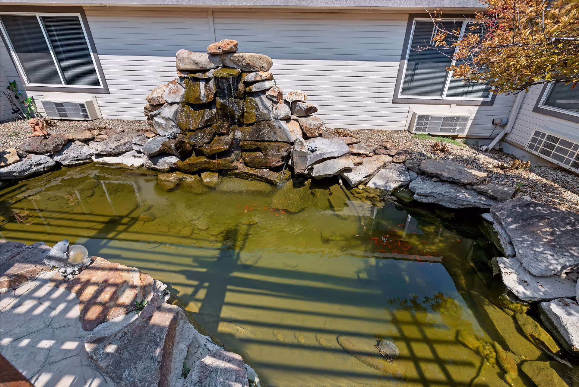 Outdoor pond with a small rock waterfall feature surrounded by large stones and gravel, adjacent to a building with white siding and windows with air conditioning units.