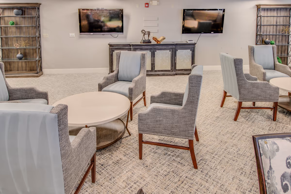A well-lit living room area with several upholstered armchairs arranged around circular coffee tables. The room features a carpeted floor, two wall-mounted flat-screen TVs, a decorative cabinet, and two wooden shelving units with decorative items.