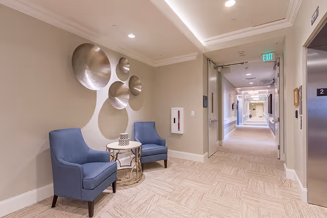 A well-lit hallway in a senior living facility with beige walls and carpeted floor. Two blue upholstered chairs are placed around a small round table with a decorative item on it. Metallic circular wall art is displayed above the chairs. The hallway extends into the distance with doors and an elevator visible on the right side.