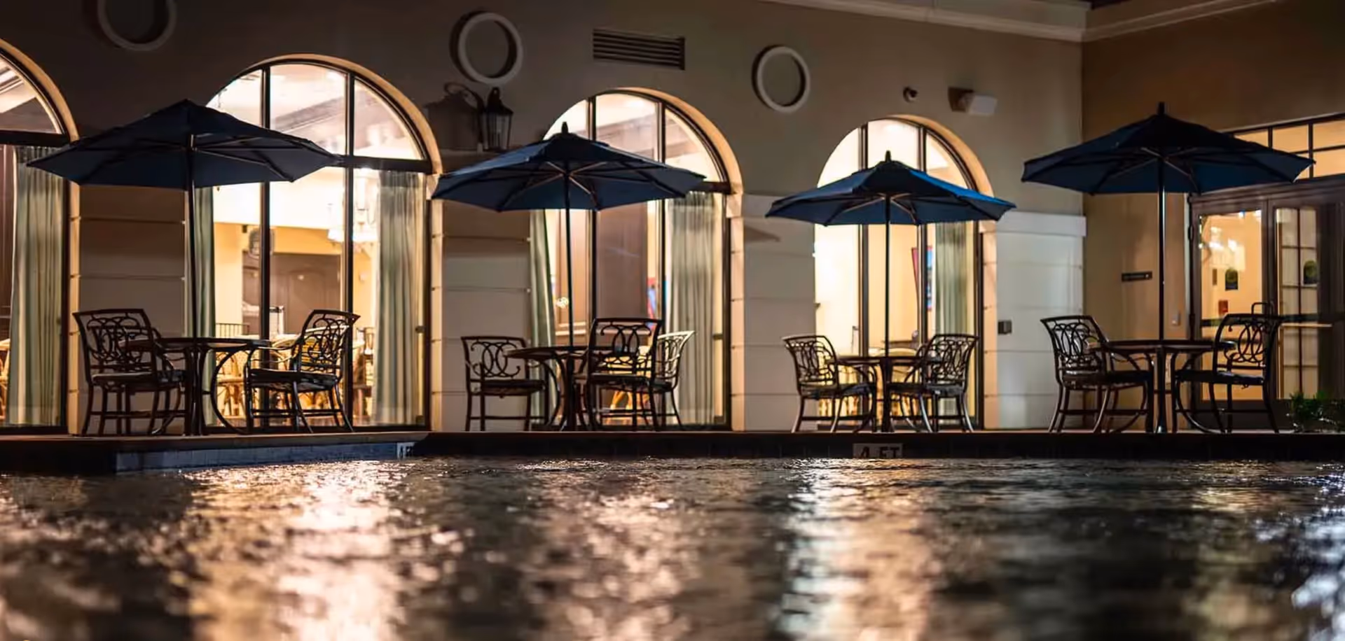 Outdoor seating area at night with several tables and chairs under large umbrellas next to a pool, with arched windows and warm interior lighting visible inside the building.