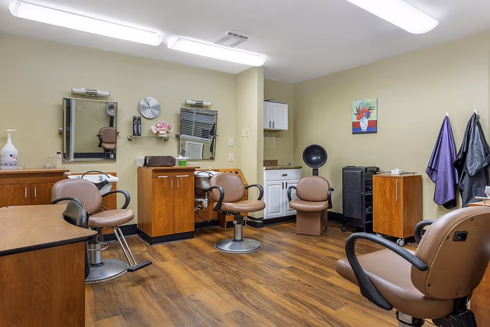 Interior view of a hair salon area in a senior living facility with multiple brown salon chairs, mirrors on the wall, wooden cabinets, a hair dryer chair, and hair styling tools. The floor is wood, and the walls are painted light yellow with a colorful painting hanging on one wall.