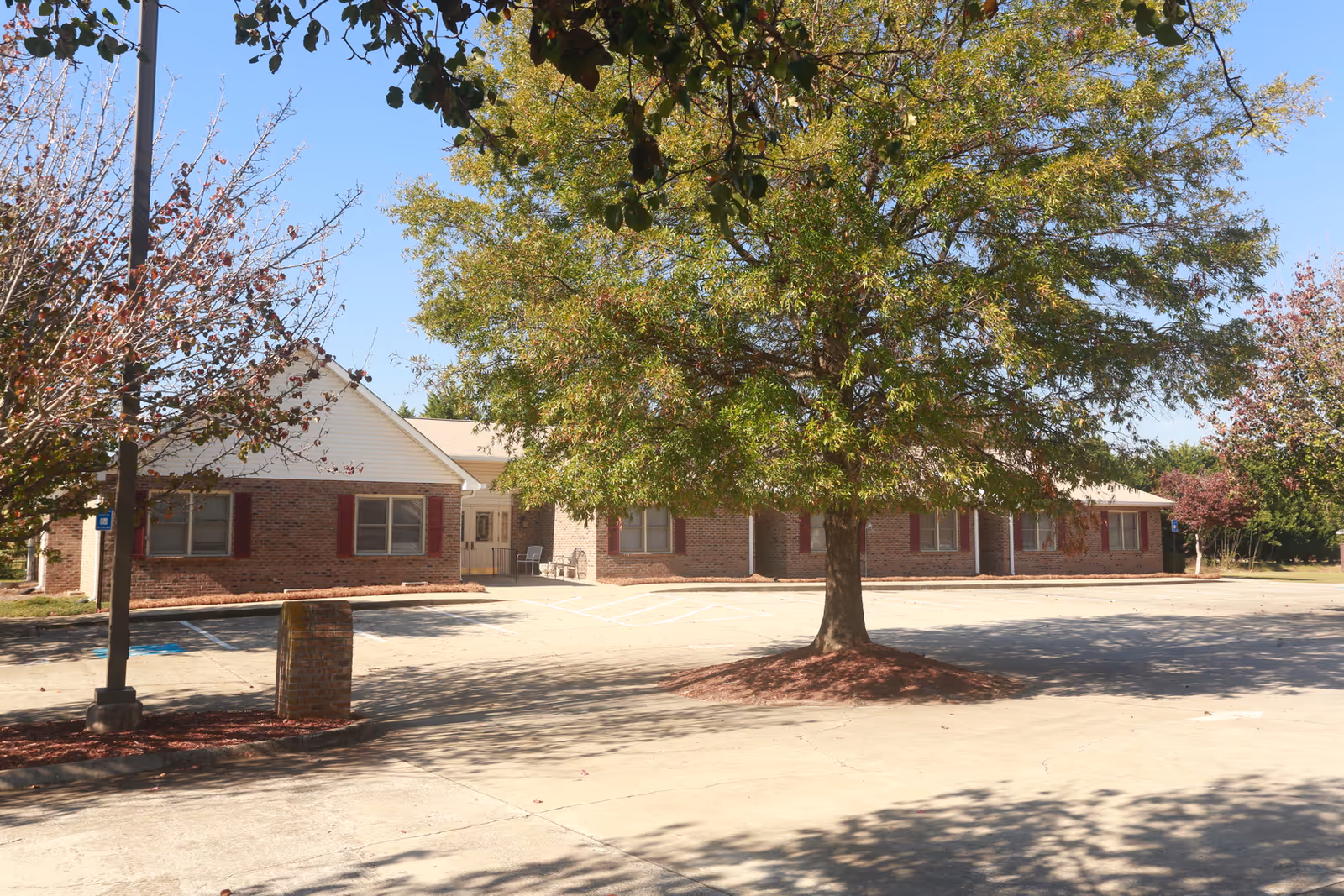 Exterior view of a single-story brick building with white siding and red shutters, surrounded by trees and a parking lot with marked spaces under a clear blue sky.