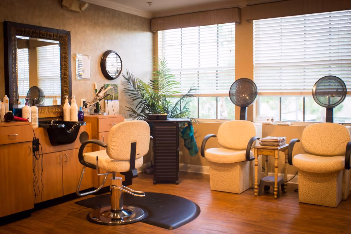 Interior of a salon area with a styling chair in front of a mirror and sink, two hair drying chairs with hooded dryers, a small table with magazines, and a large window with blinds letting in natural light.