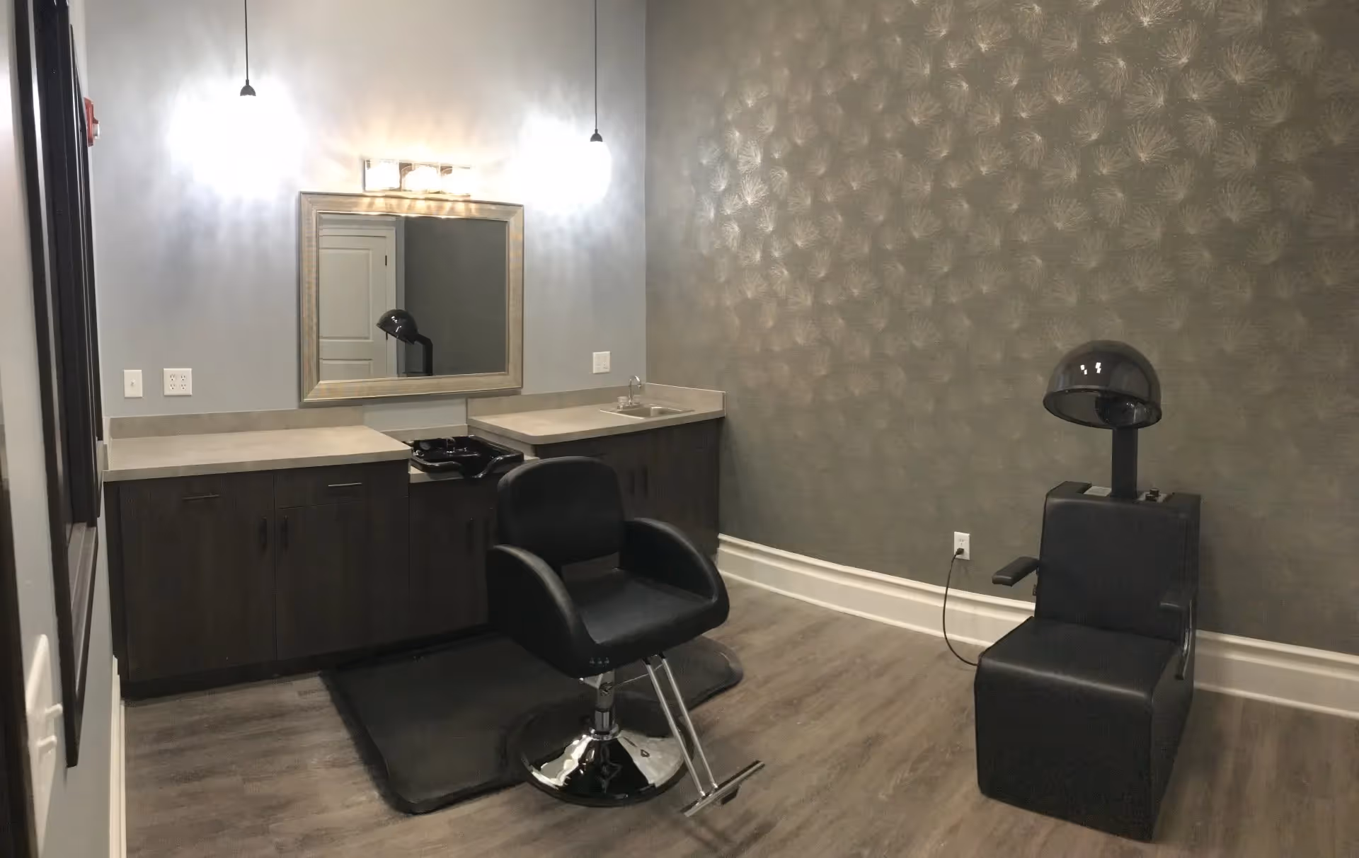Interior of a salon room with a black salon chair in front of a counter with a mirror and two hanging lights. To the right, there is a black hair dryer chair against a textured wall.