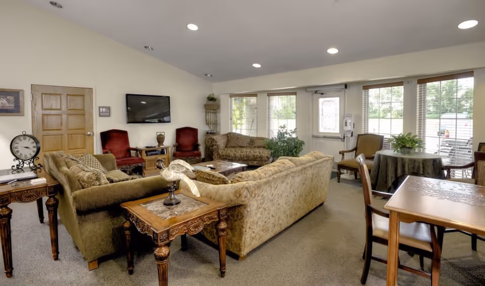 A cozy living room area in a senior living facility with multiple upholstered sofas and chairs arranged around a central coffee table. There is a flat-screen TV mounted on the wall, a wooden side table with a decorative clock, and large windows letting in natural light. A round table with a green tablecloth and chairs is visible near the windows, along with some potted plants.